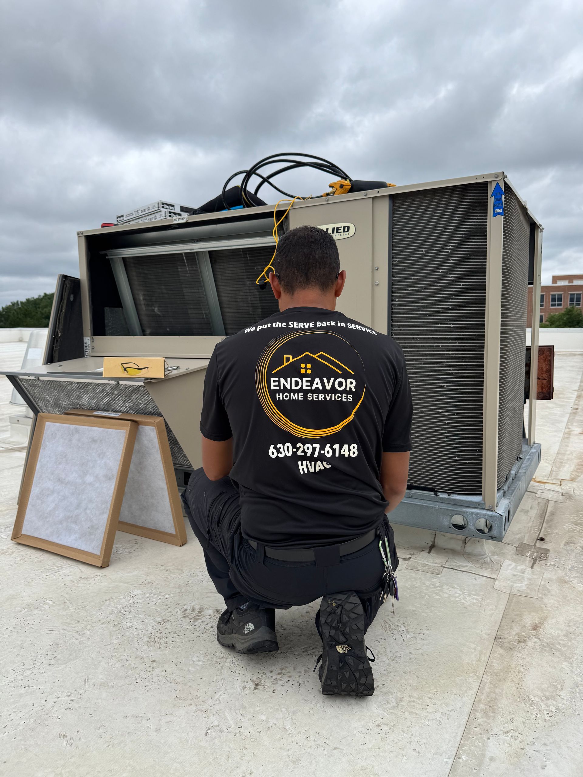 HVAC technician kneeling in front of an AC unit on a rooftop, wearing a black shirt with company logo. Cloudy sky.