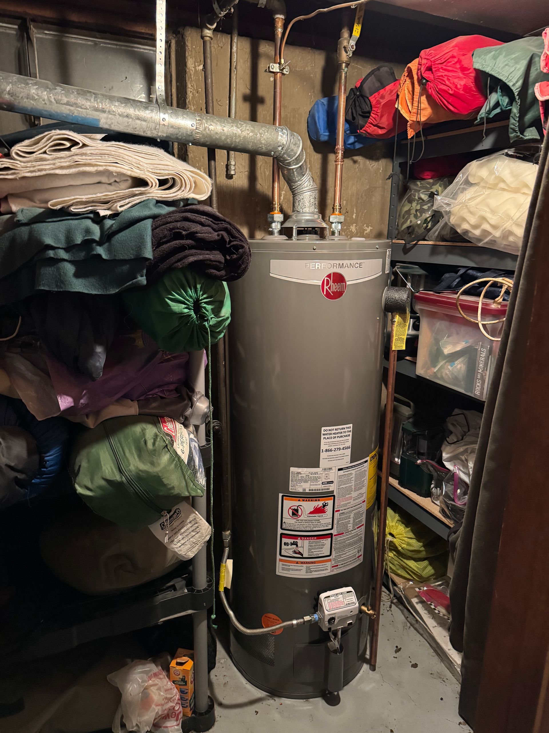 Water heater in a cluttered basement with shelving and stored items.
