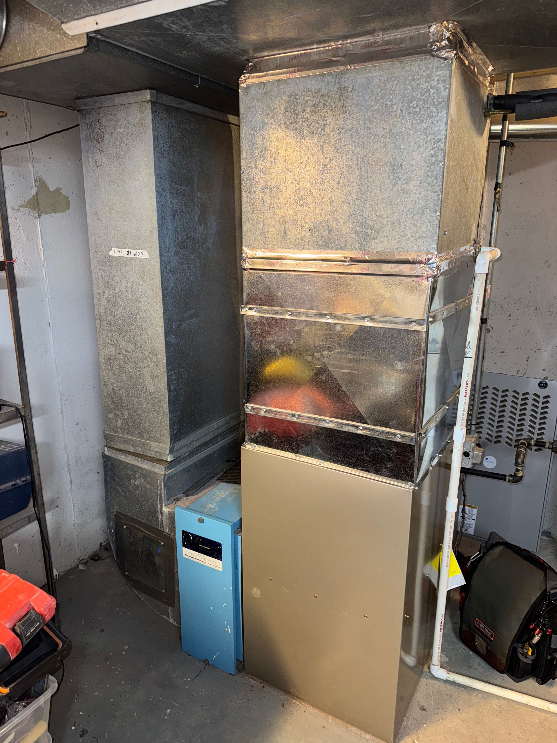 Furnace and ductwork in a basement setting. Metal components: silver, blue, tan.  Near a concrete wall.