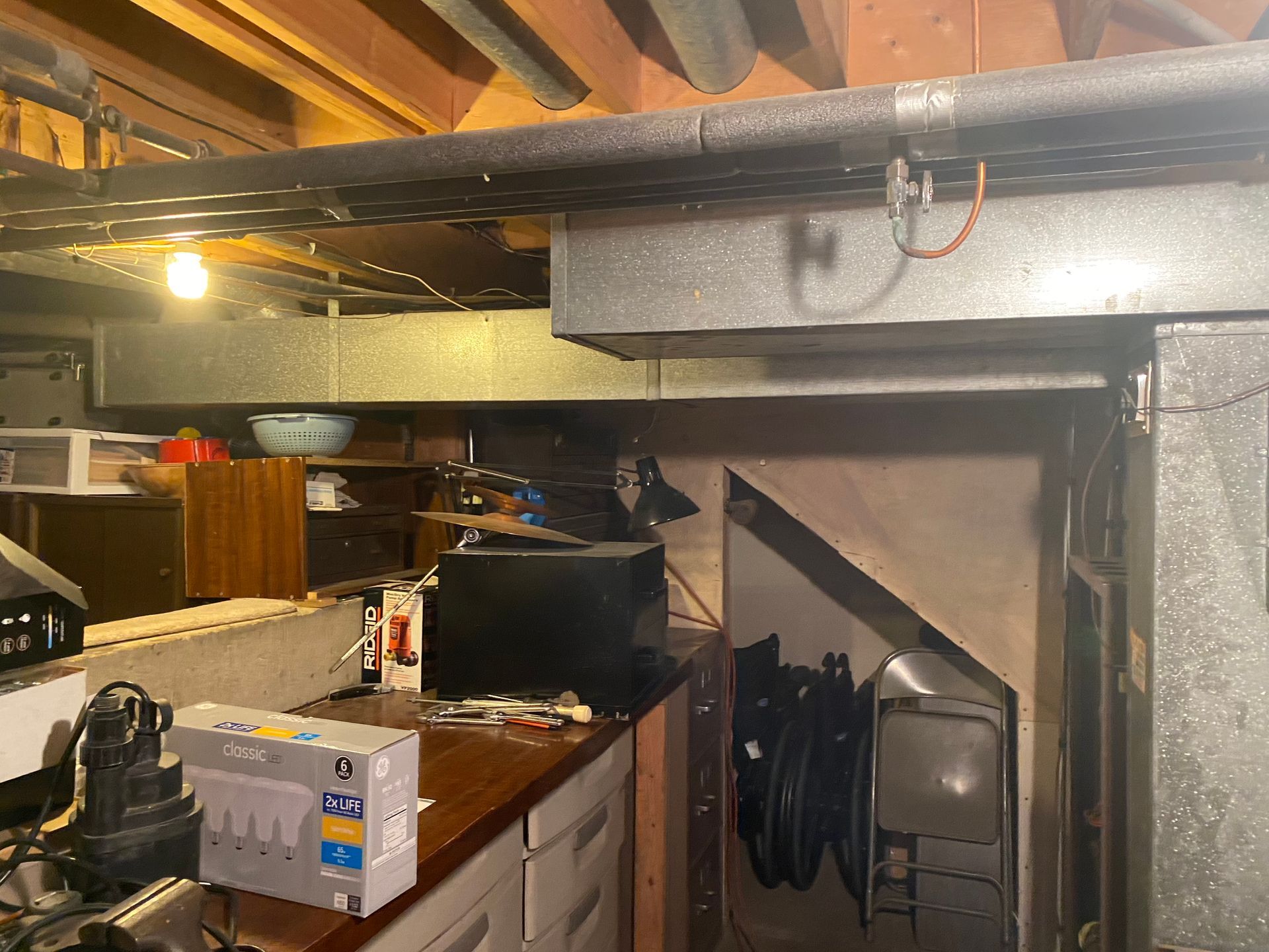 Basement storage area with workbench, shelving, and old TV. Ductwork and exposed pipes overhead.