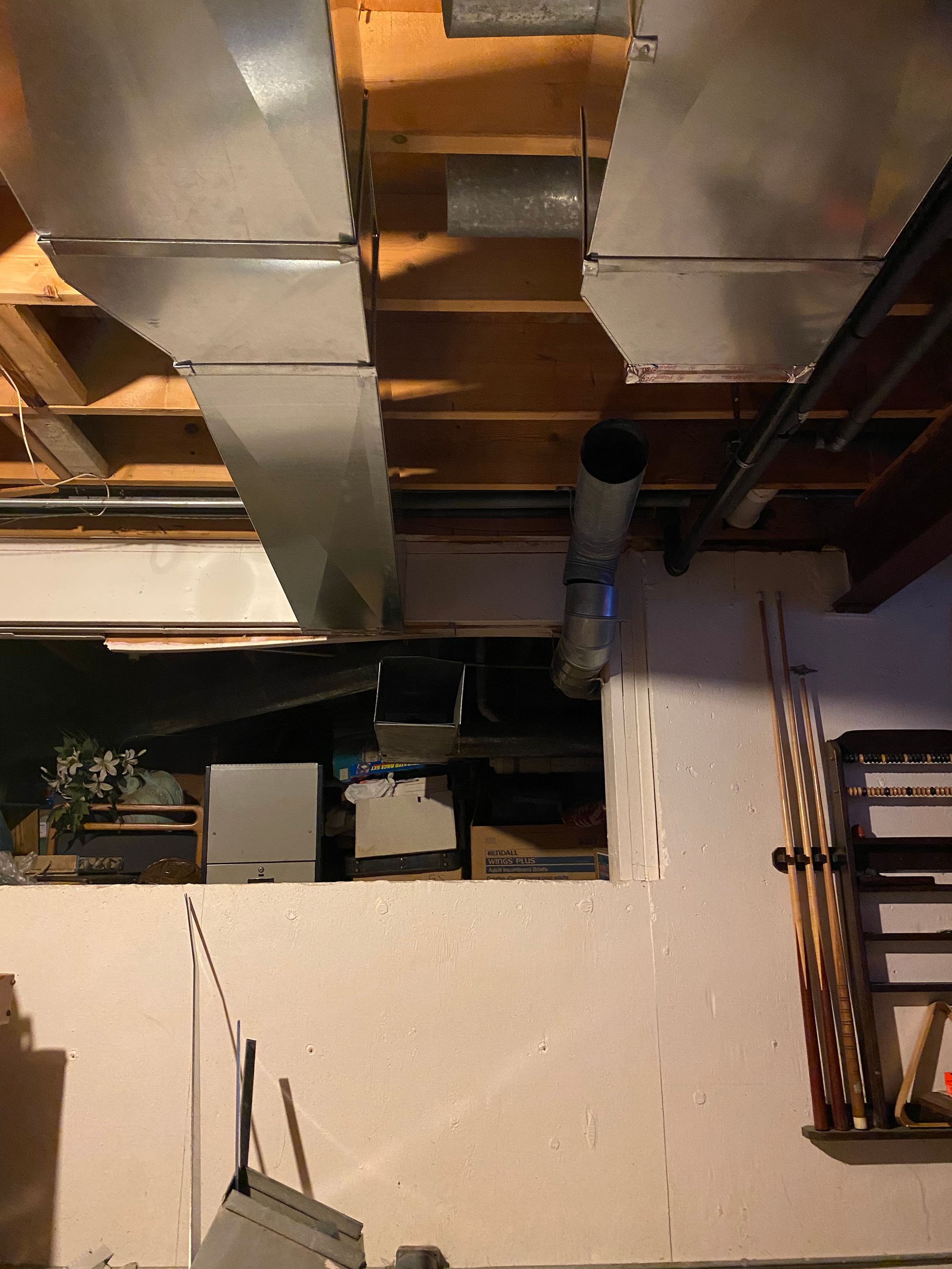 Basement ceiling with metal ducts, a vent pipe, and exposed wooden beams above a concrete wall.