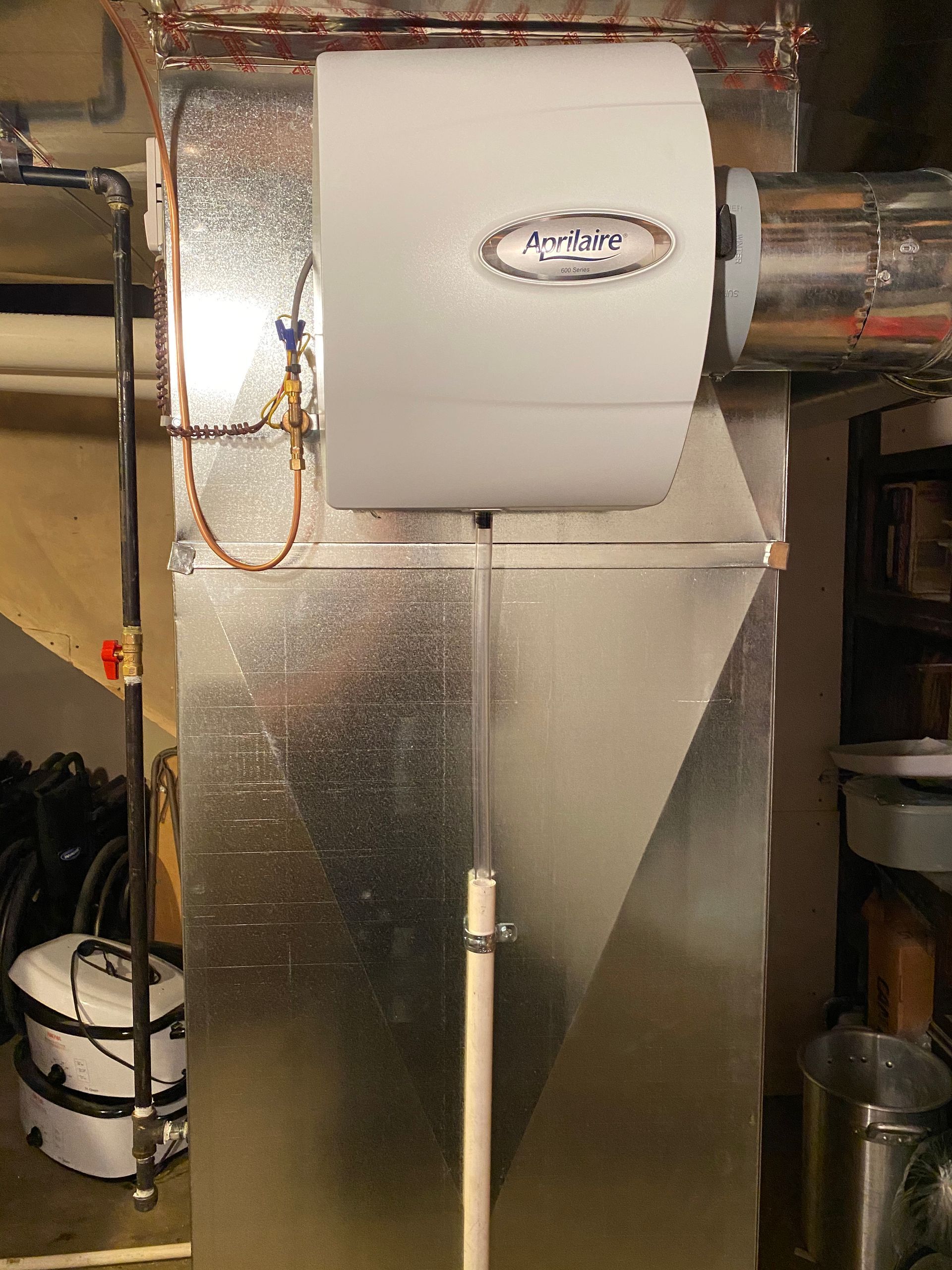 Humidifier attached to HVAC ductwork in a basement. White unit with a drainpipe and water line.