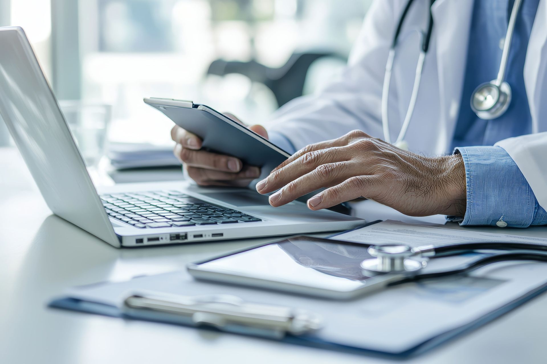 Um médico vestindo um jaleco branco e um estetoscópio digita em um laptop enquanto segura um tablet em uma mesa.