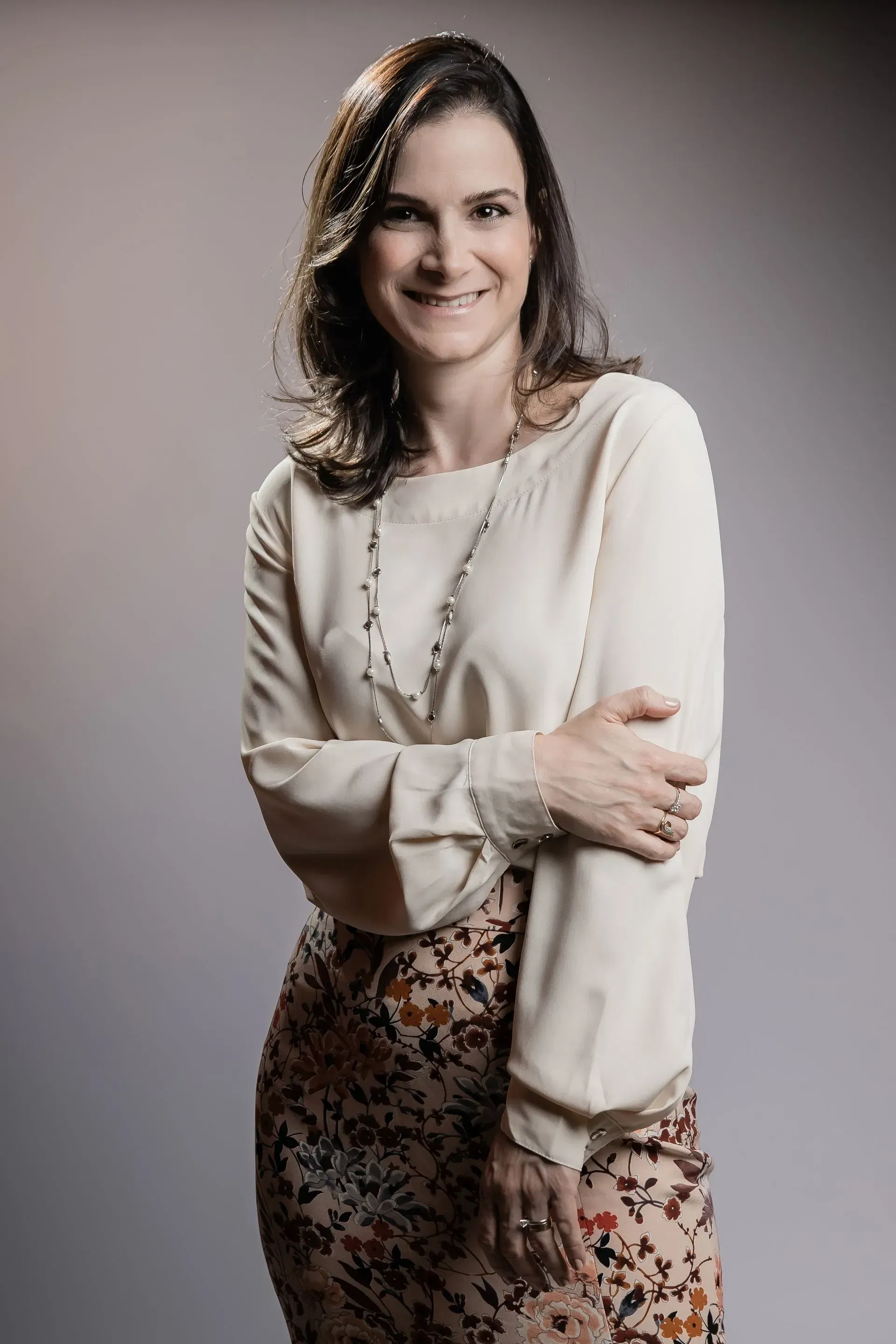 Uma mulher de camisa branca e saia florida está de pé com os braços cruzados e sorrindo.