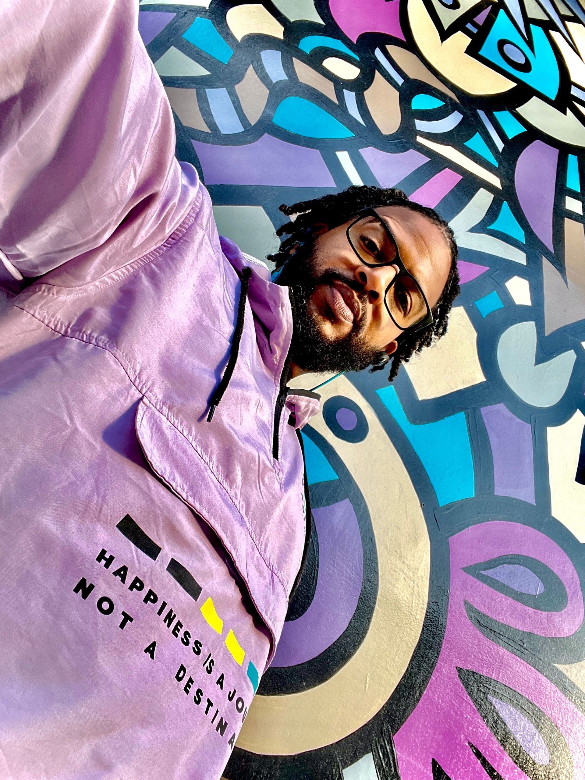 Antjuan wearing glasses and a purple jacket is laying in front of a colorful wall