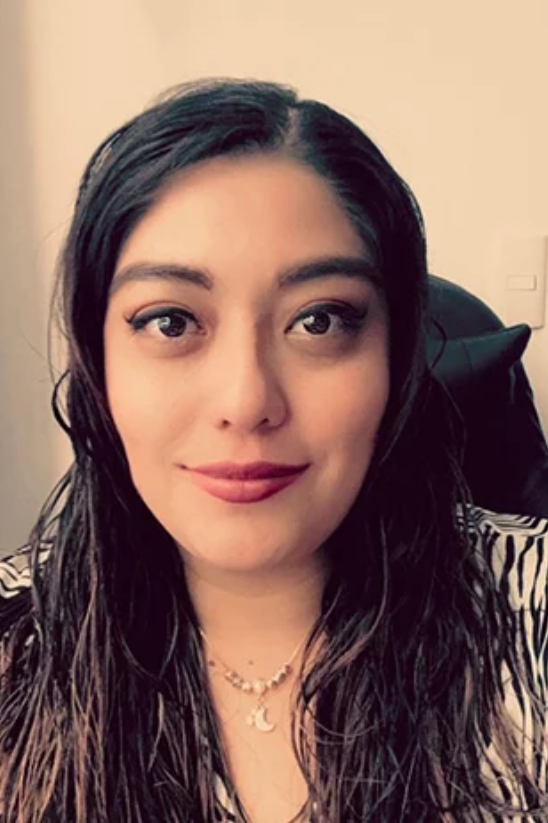 Una mujer con cabello largo y oscuro, con un top estampado y un collar, sonríe a la cámara.