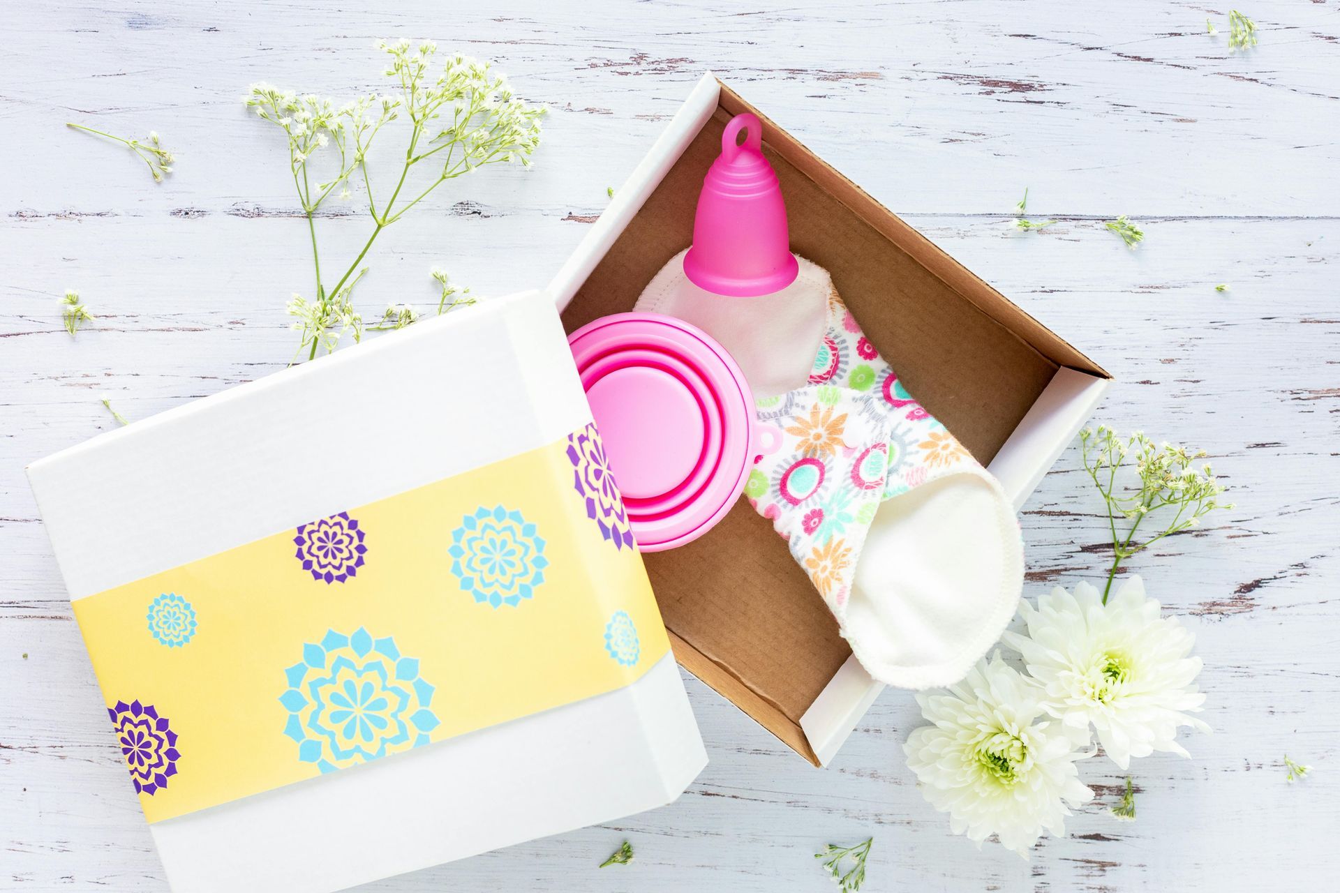 Caja de regalo abierta con copa menstrual, compresa de tela y disco limpiador. Flores blancas y un lazo amarillo adornan la caja.