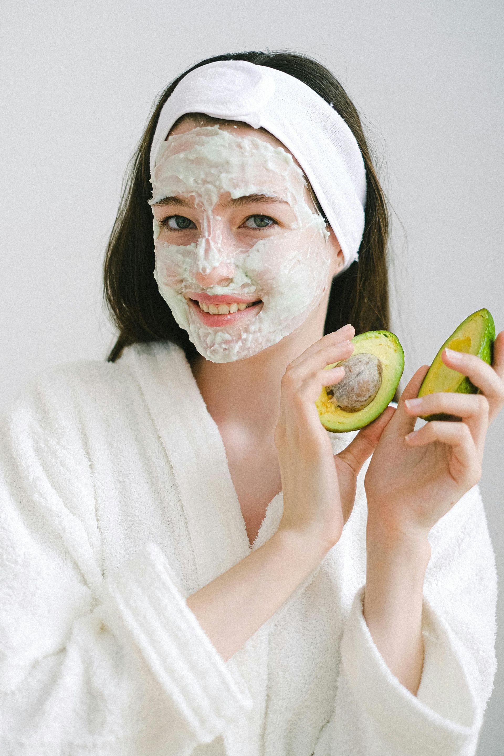 Mujer en bata con mascarilla, sosteniendo mitades de aguacate y sonriendo. Lleva una diadema blanca.