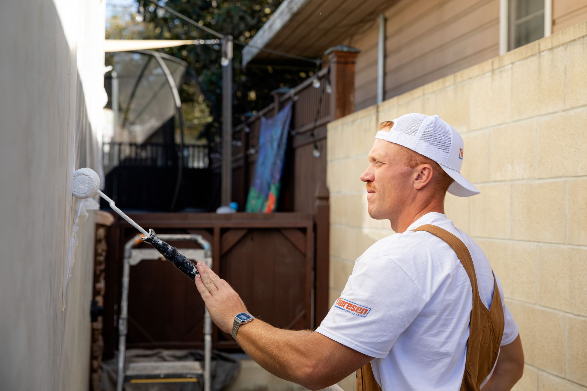 Man painting a wall with a roller outdoors. He wears overalls and a white hat.