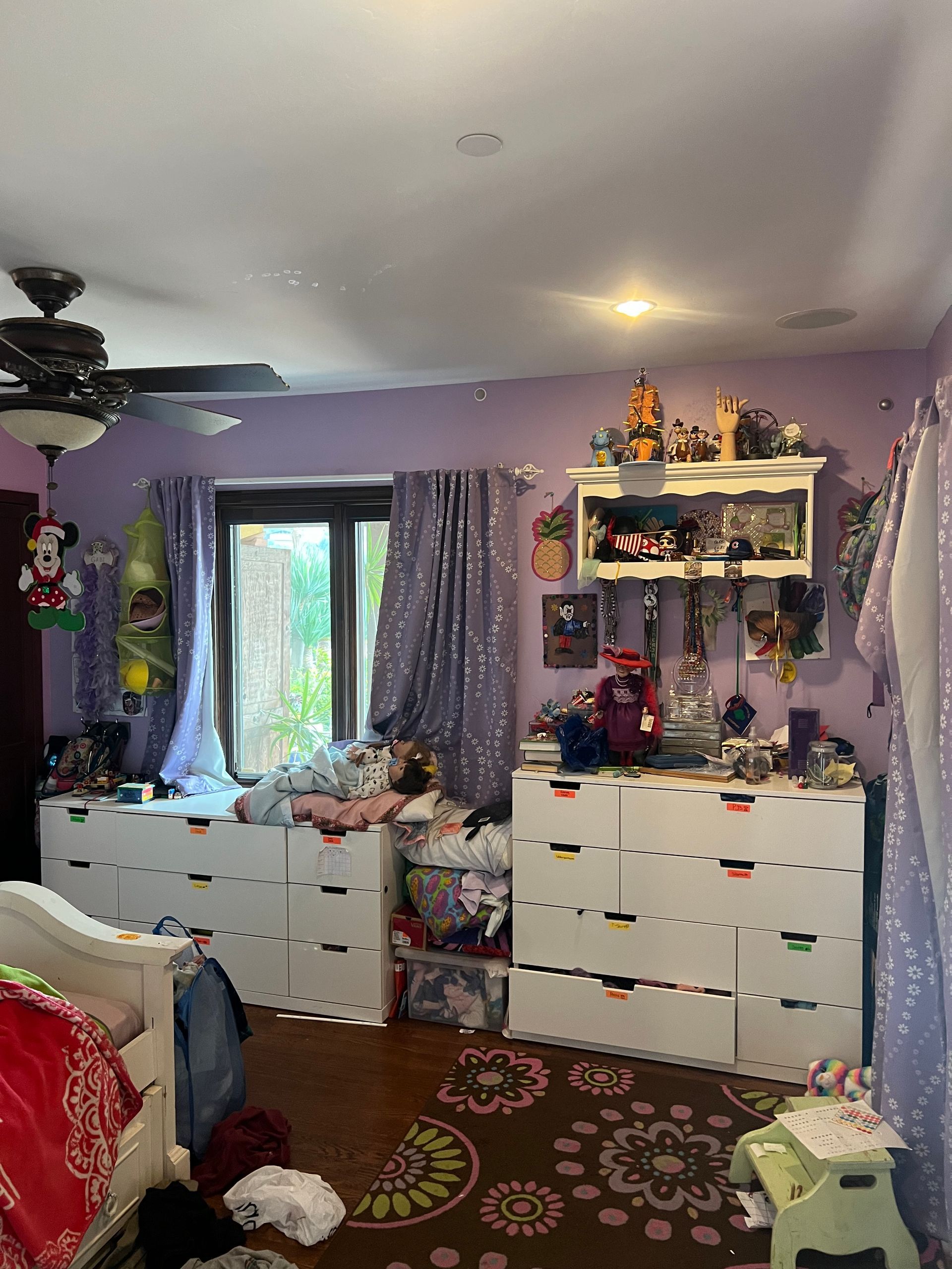 A bedroom with purple walls , white dressers , and a ceiling fan.
