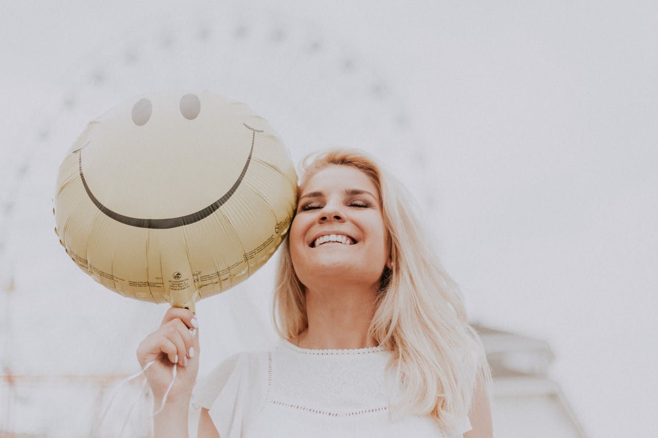 A woman is holding a smiley face balloon to the right of her face.