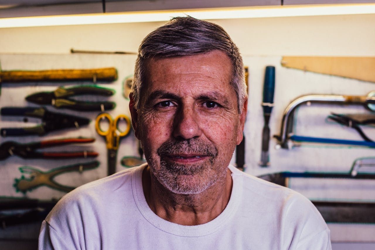 A man with a beard is sitting in front of a wall with tools on it.