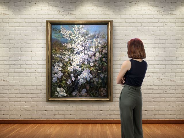 A person stands in a gallery, viewing a large framed painting of white flowers against a white brick wall.