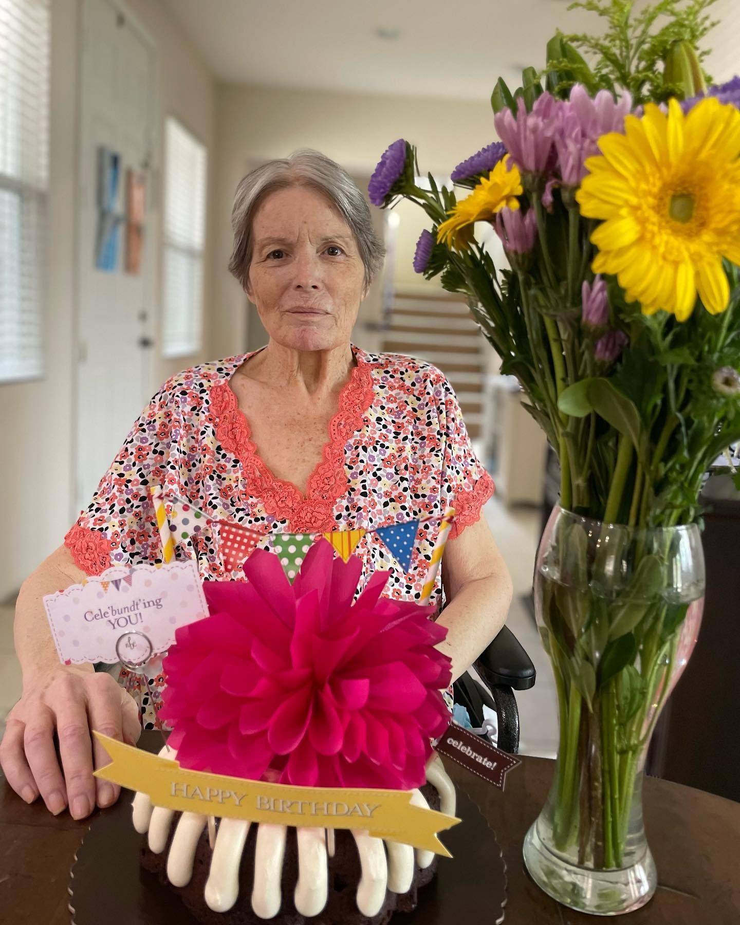 A woman in a wheelchair is sitting at a table with a birthday cake and flowers.