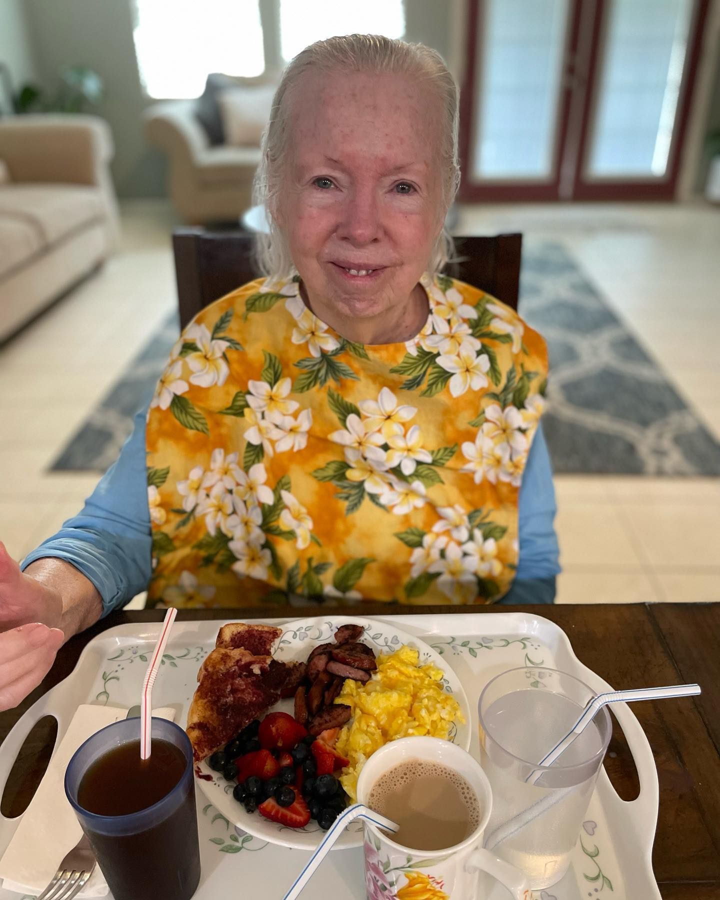 An elderly woman wearing a bib is sitting at a table eating breakfast.