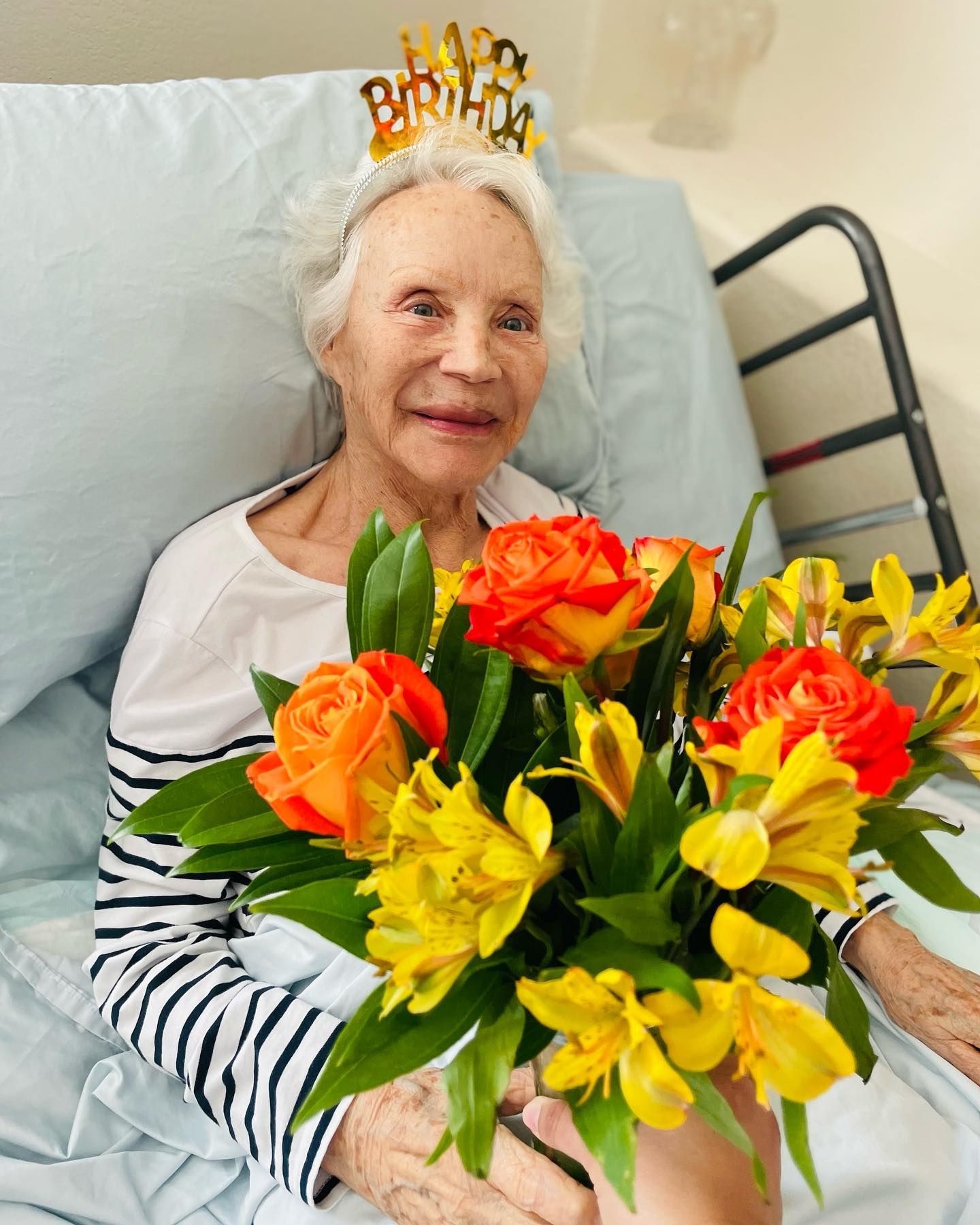 An elderly woman is sitting in a hospital bed holding a bouquet of flowers.