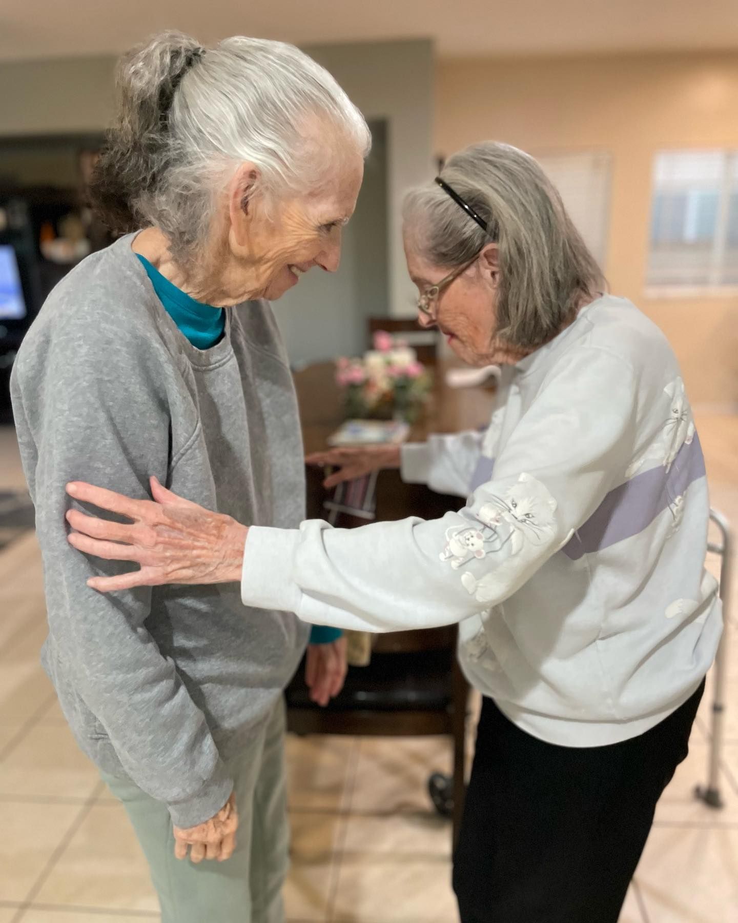 Two older women are hugging each other in a living room.