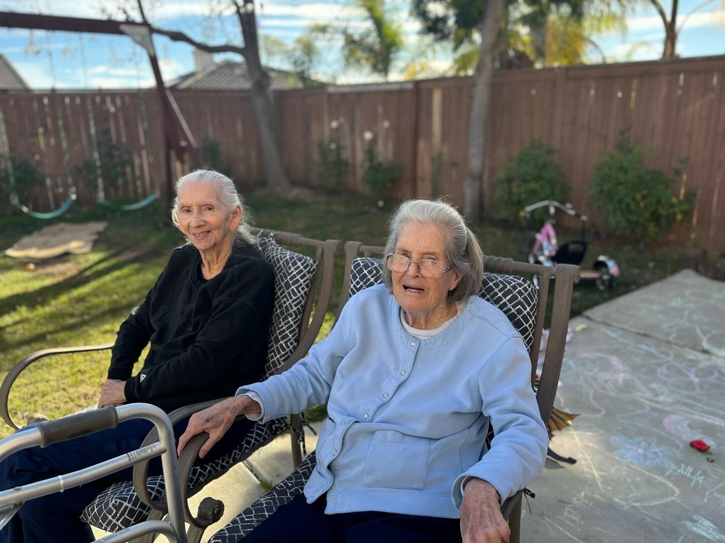 Two elderly women are sitting on a swing in a backyard.