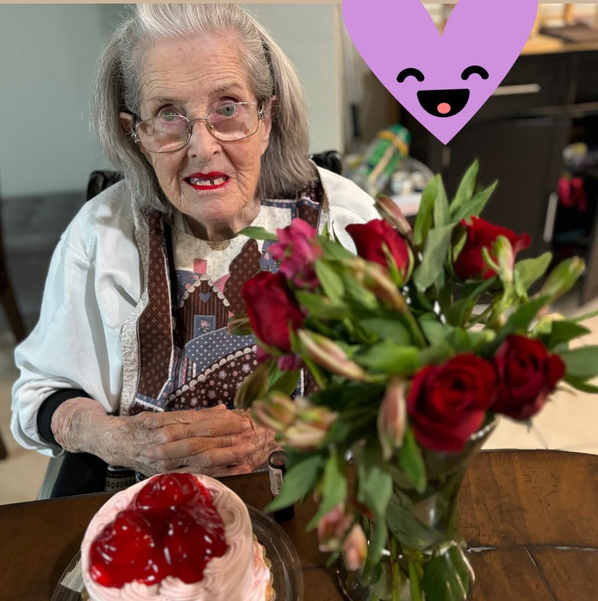 An elderly woman sits at a table with flowers and a cake