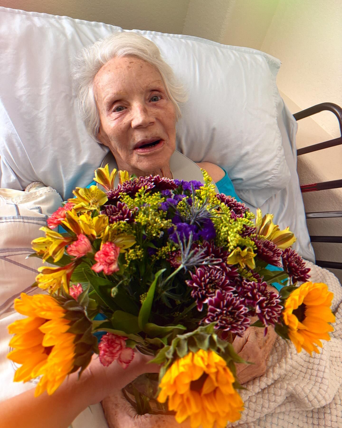 An elderly woman in a hospital bed is holding a bouquet of flowers