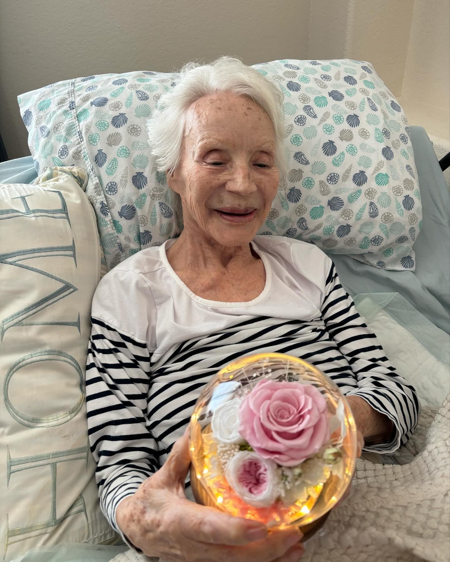 An elderly woman is sitting in a bed holding a glass ball with a pink rose in it.