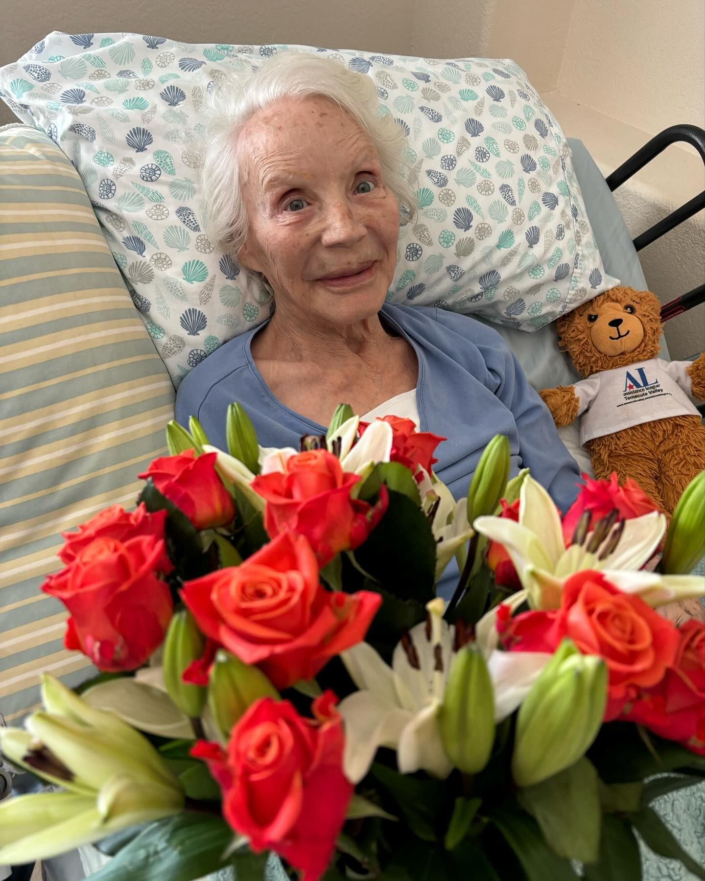 An elderly woman is laying in a hospital bed with a bouquet of flowers and a teddy bear.