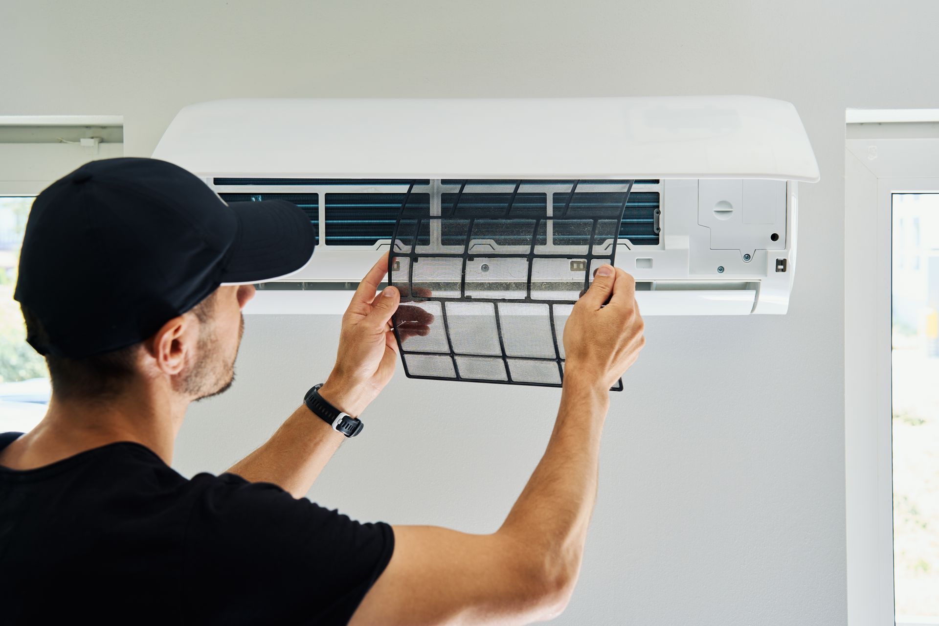 Man in black cap removing an air filter from a white wall-mounted air conditioner.