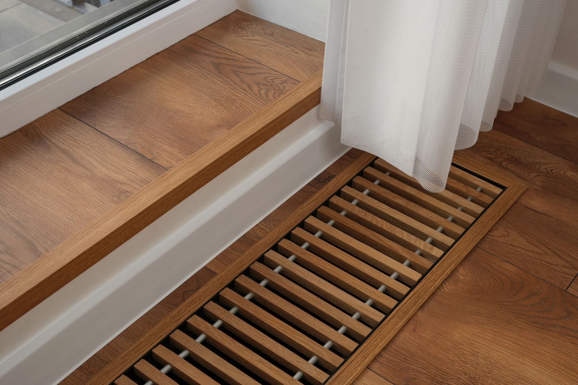 Wooden floor with a window ledge, a ventilation grill, and a white curtain.