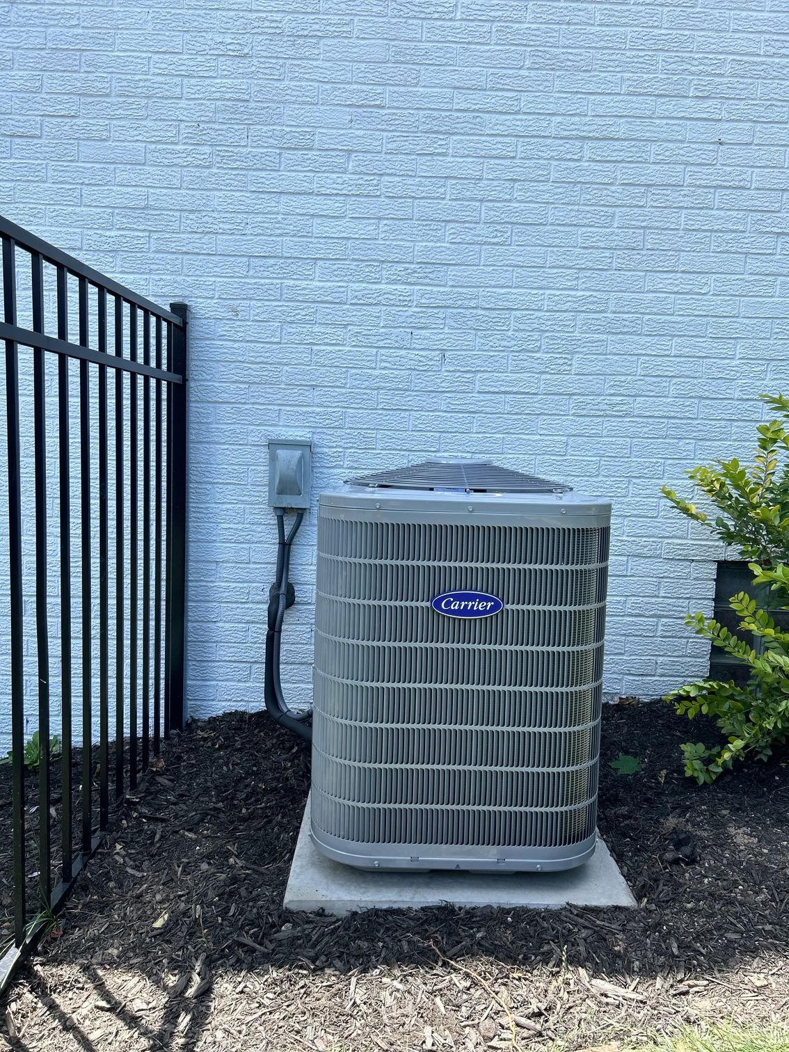 A carrier air conditioner is sitting outside of a brick building next to a fence.