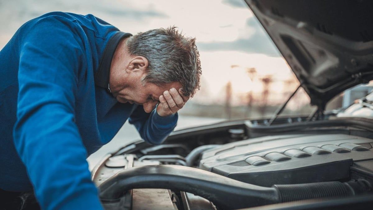 Man with hand on forehead, distressed, looking at car engine with open hood.