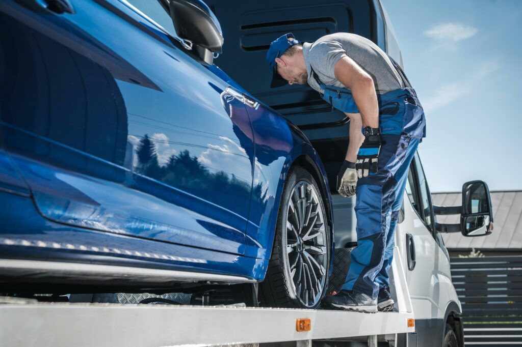 A tow truck operator in blue overalls prepares to secure a blue car on a flatbed tow truck.