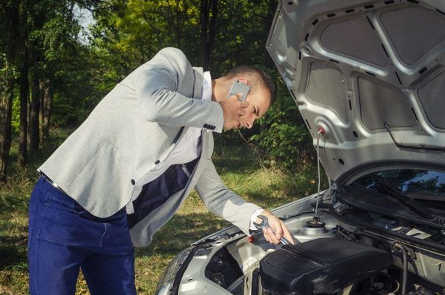 Man in suit on phone, looking at car engine, hood open, outdoors.