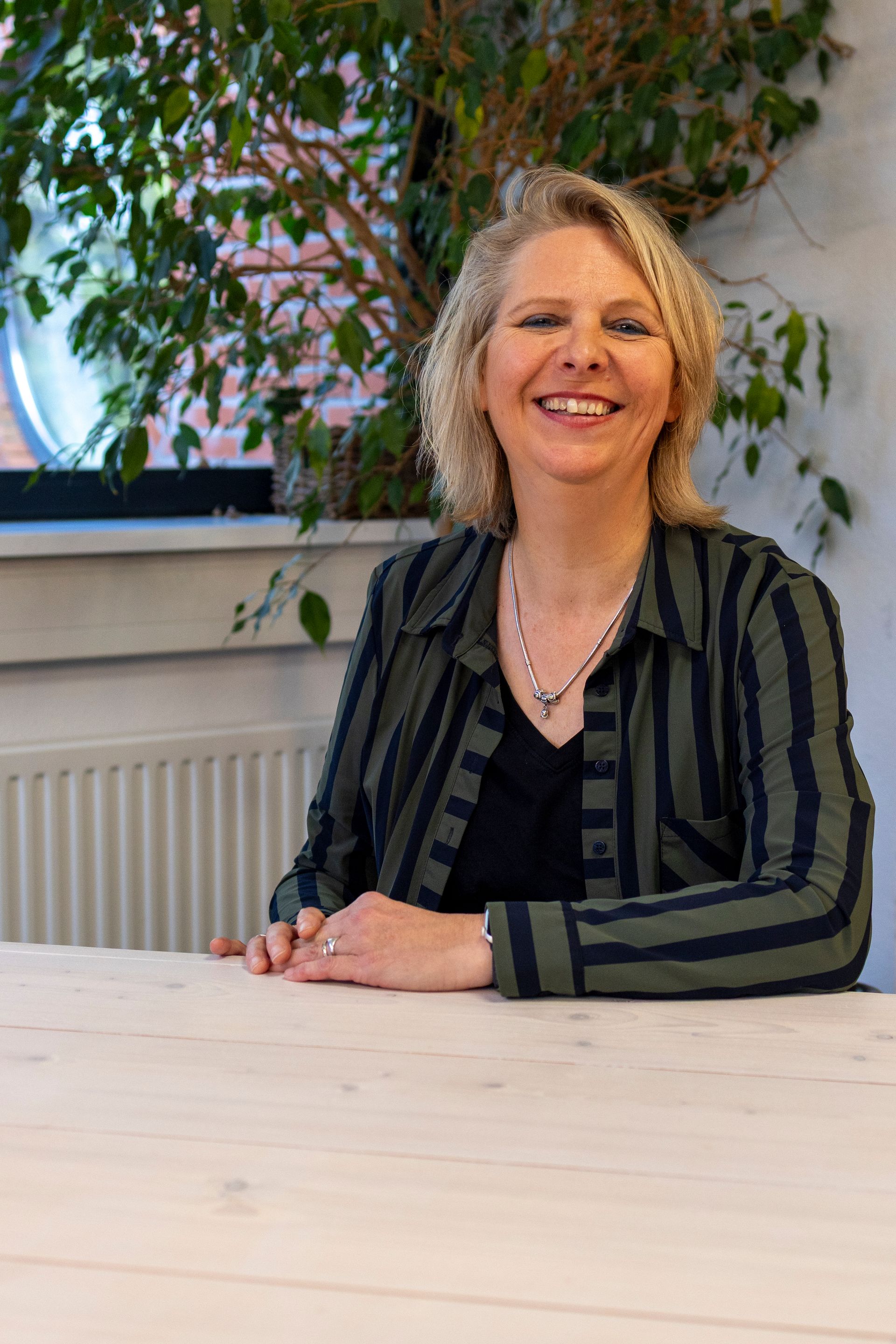 Vrouw glimlacht aan een tafel, gekleed in een gestreepte jas. Binnenomgeving met een plant op de achtergrond.