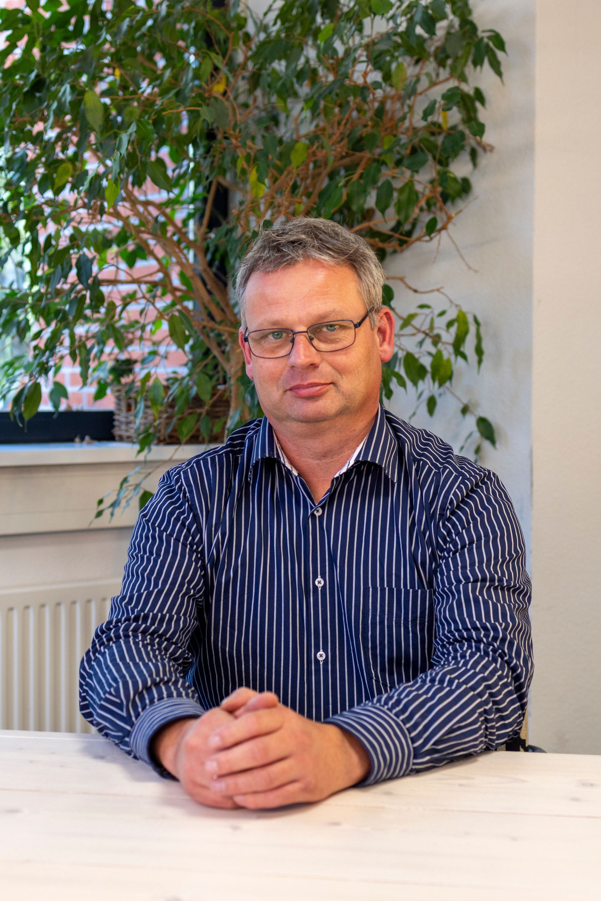 Een man met een bril en een blauw gestreept shirt zit met zijn handen ineengevouwen aan een tafel, omringd door groen.