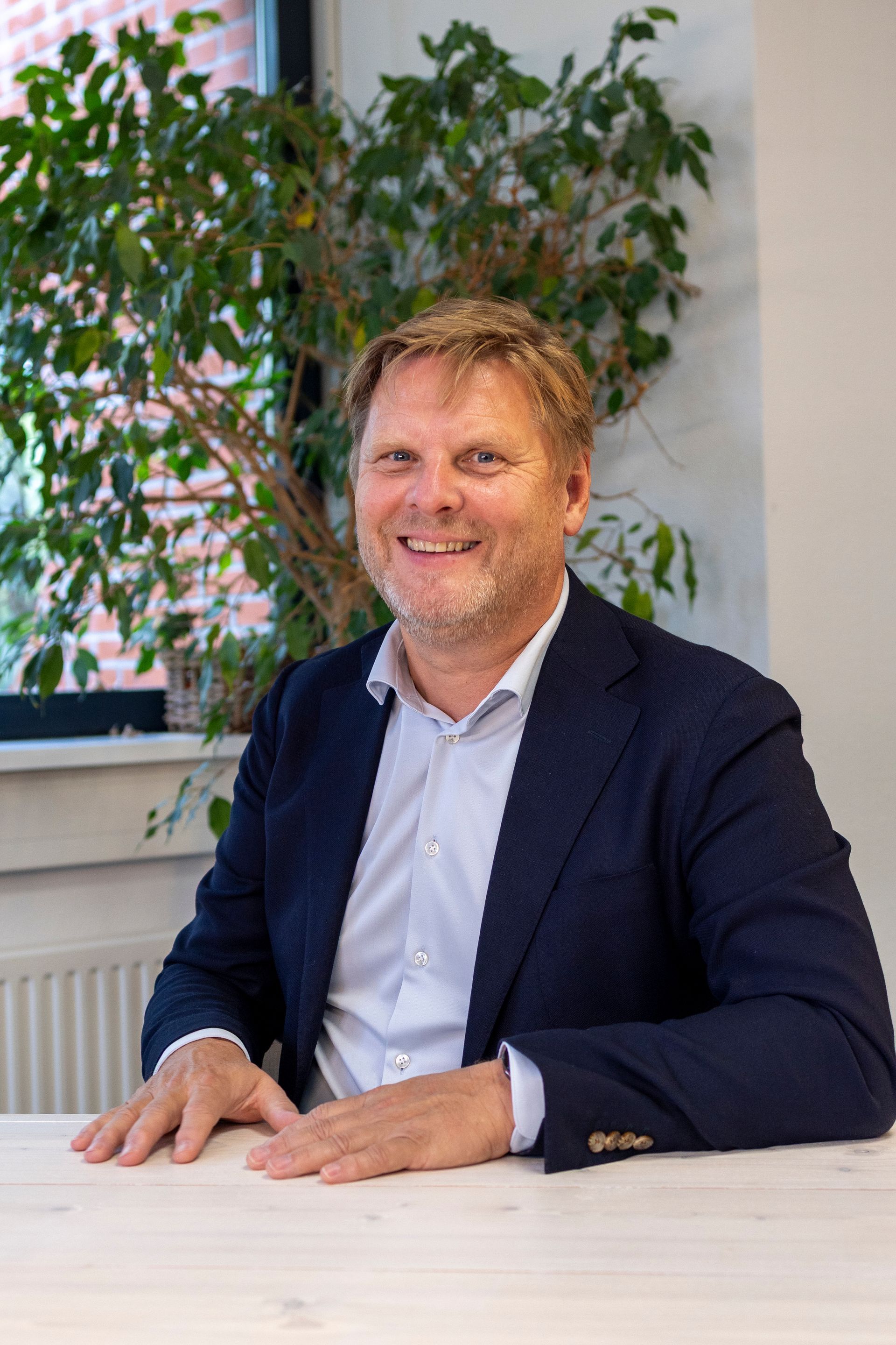 Een man in een blazer glimlacht achter een bureau, in een binnenruimte met groen op de achtergrond.