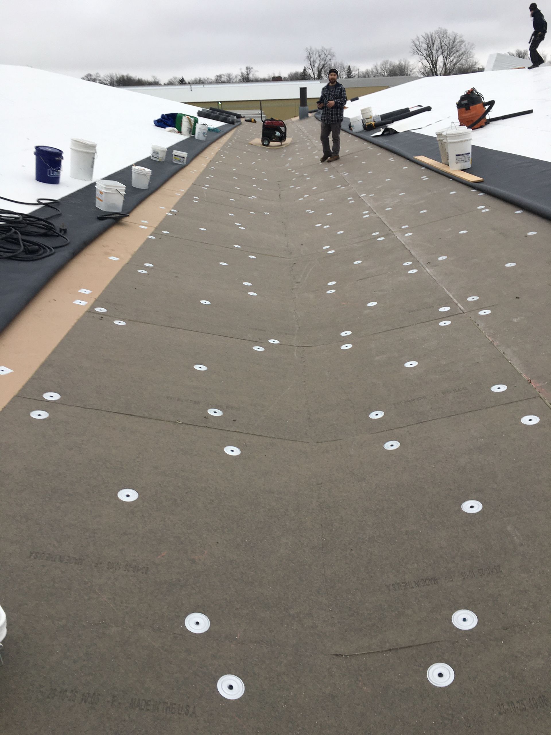Workers on a roof, installing roofing material with multiple white circular fasteners.