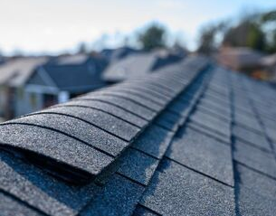 Close-up of a dark asphalt shingle roof, with a blurred background of houses on a sunny day.