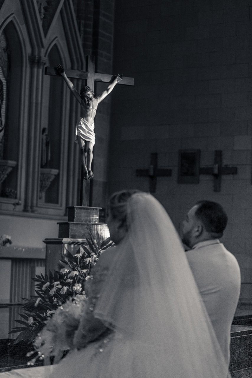 Una novia y un novio están arrodillados frente a una cruz en una iglesia.
