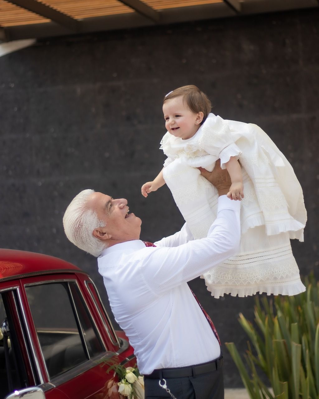 Un hombre sostiene a un bebé en sus brazos delante de un coche rojo.