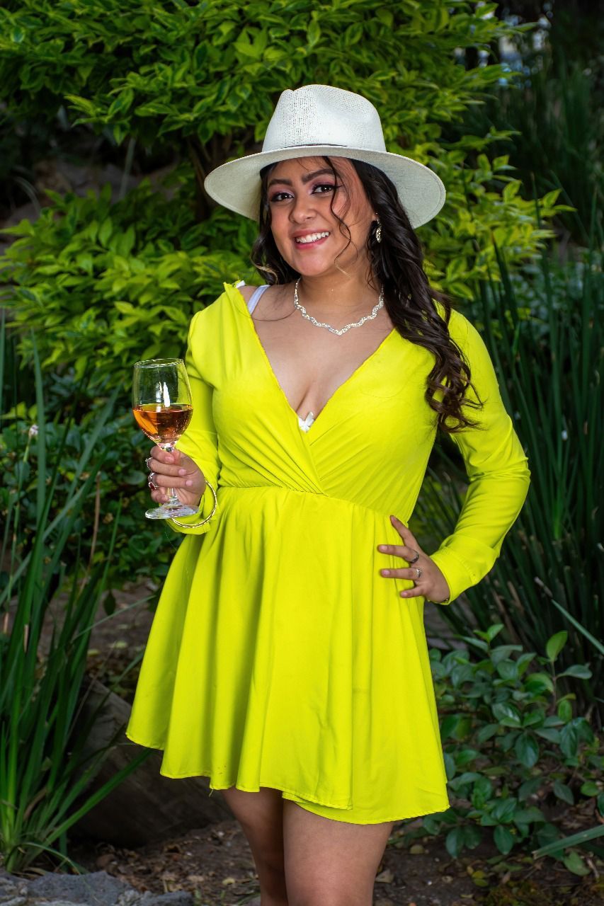 Una mujer con un vestido amarillo y un sombrero blanco sostiene una copa de vino.