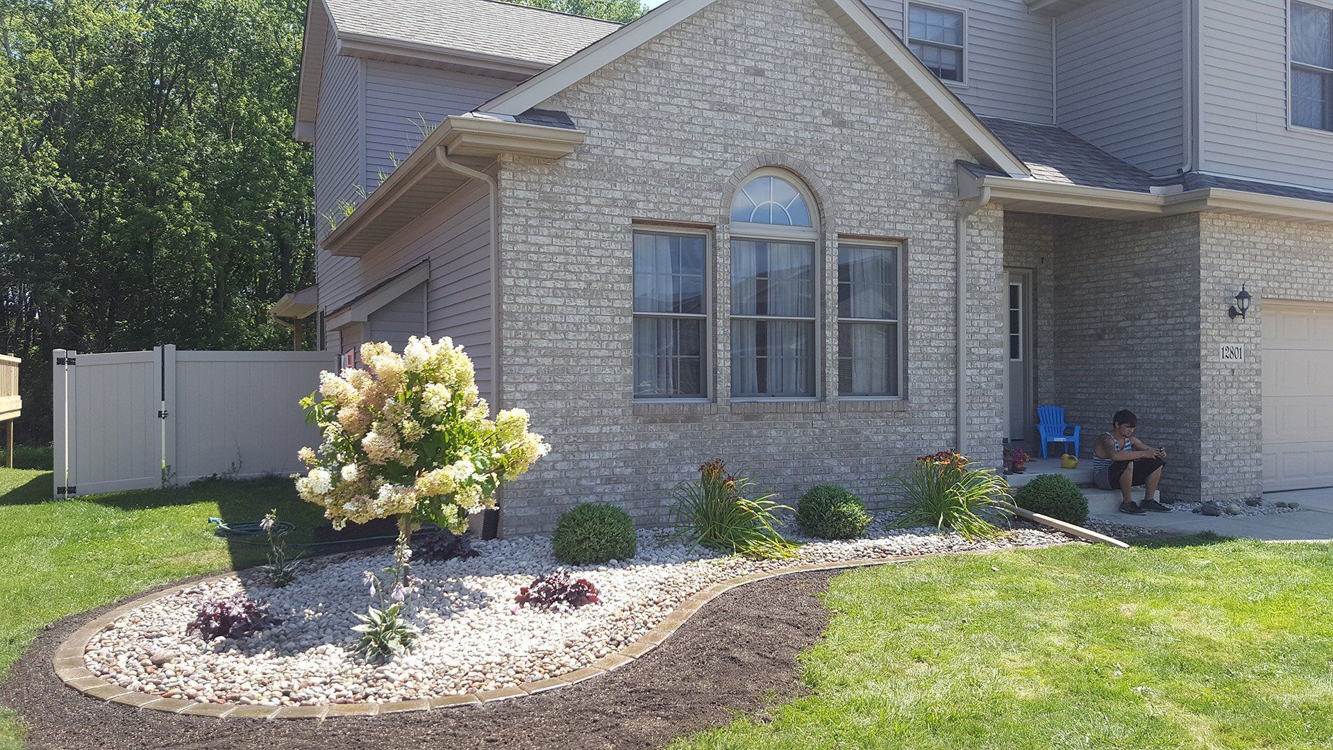 A house with a brick facade and landscaping, including a curved flower bed with white stones and blooming plants.