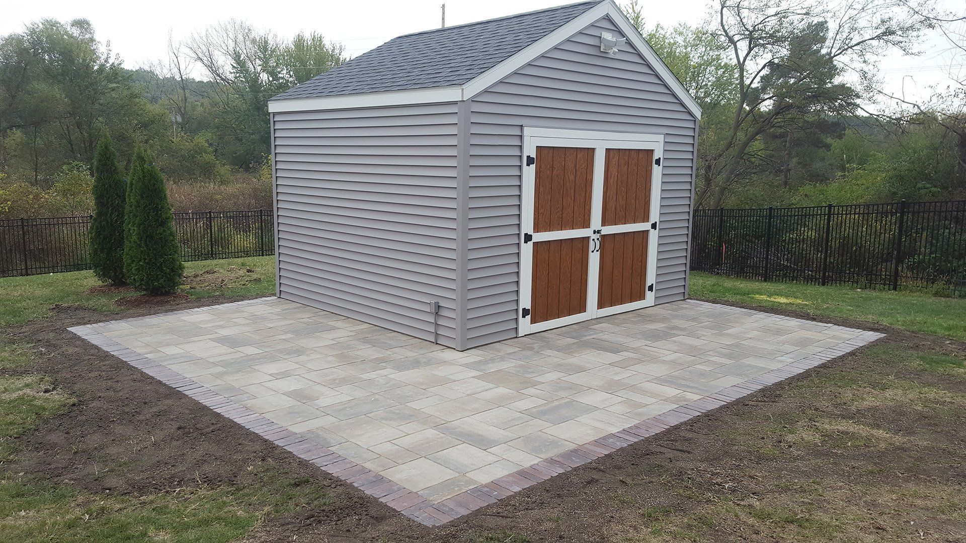 Gray shed with brown doors on a paved patio in a grassy backyard.