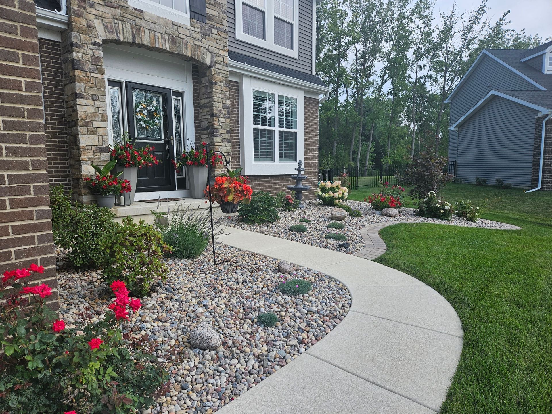A curved concrete walkway leads to a house with a stone and brick facade, surrounded by a rock garden and colorful flowers.