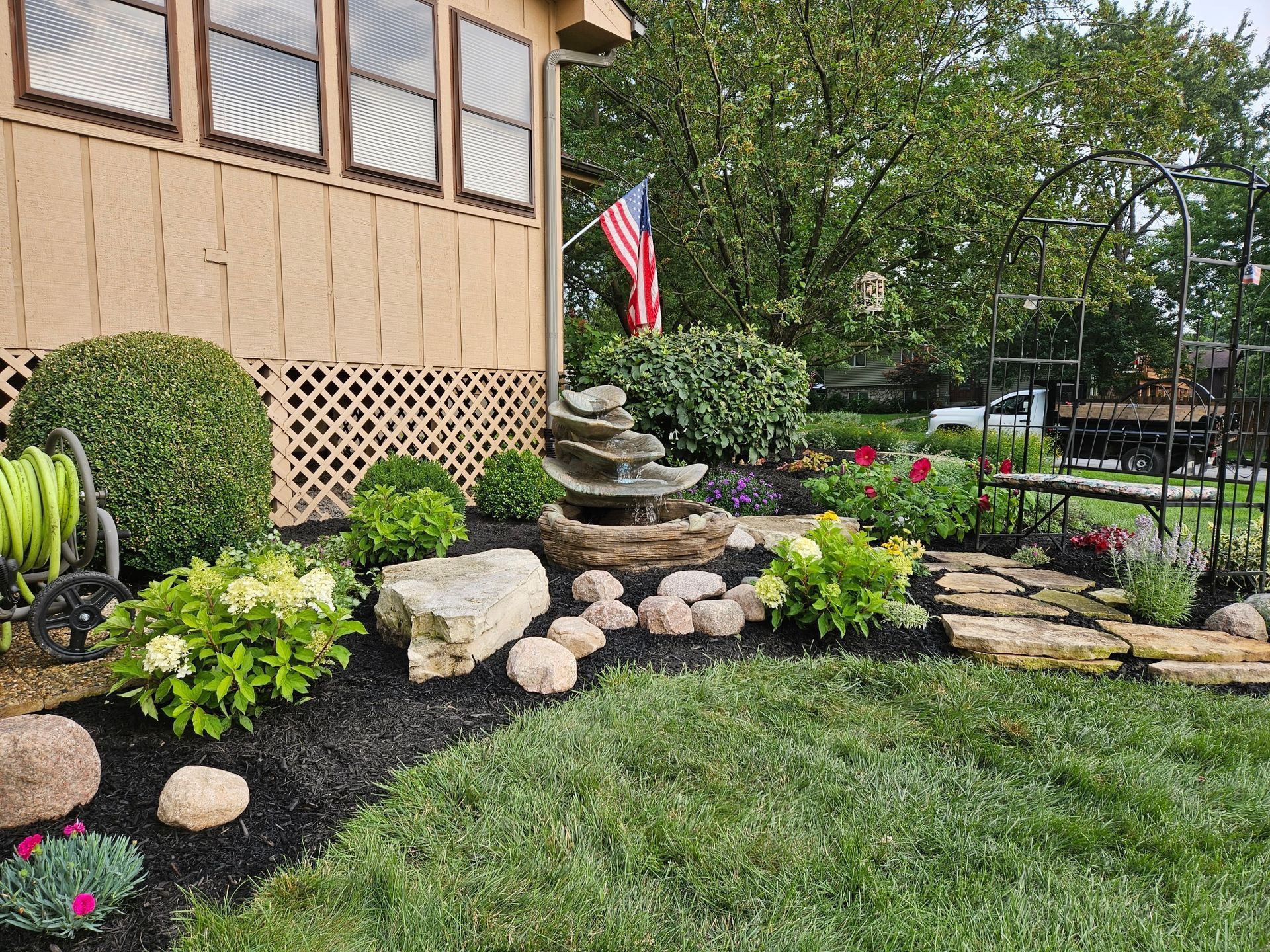 Well-landscaped yard with water fountain, flowers, and American flag in front of a tan house.