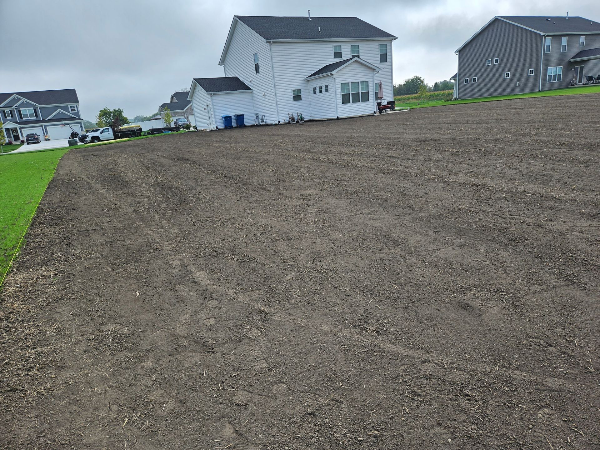 Graded dirt field in front of two-story houses; cloudy sky; green grass on the left.