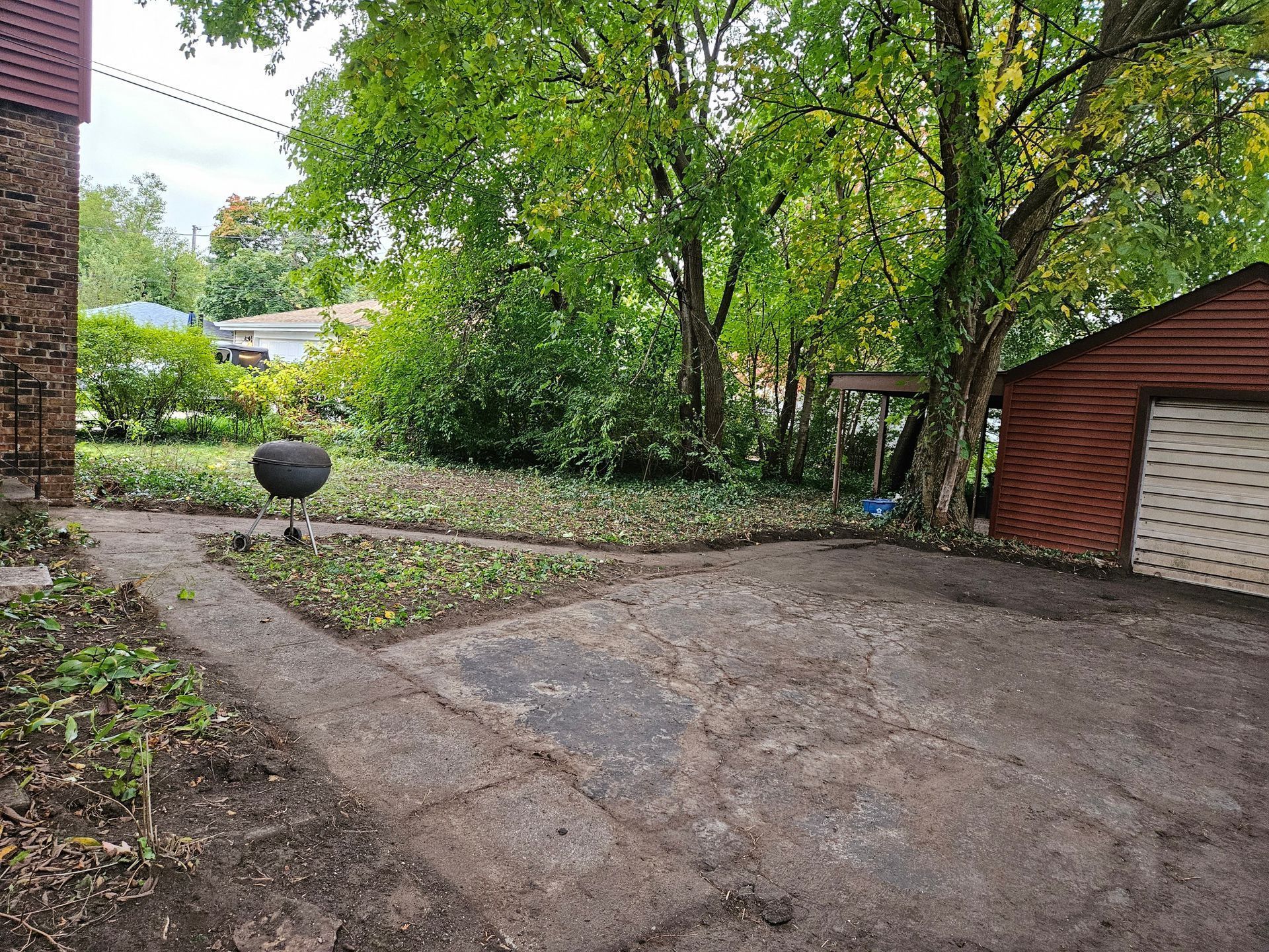 Concrete driveway and yard with trimmed garden beds, black grill, and trees. Red garage at right.