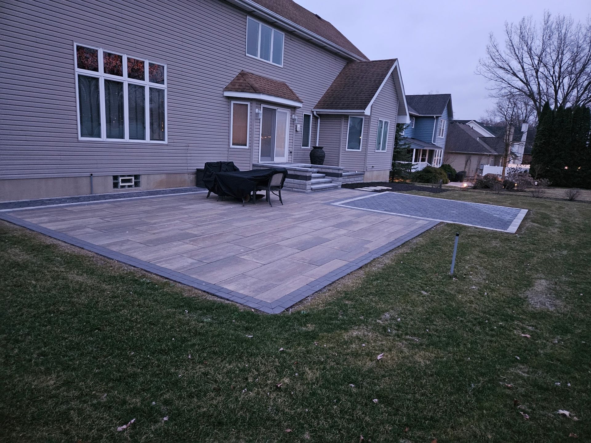 Backyard patio with gray pavers and house with dark siding. Grassy lawn surrounds.