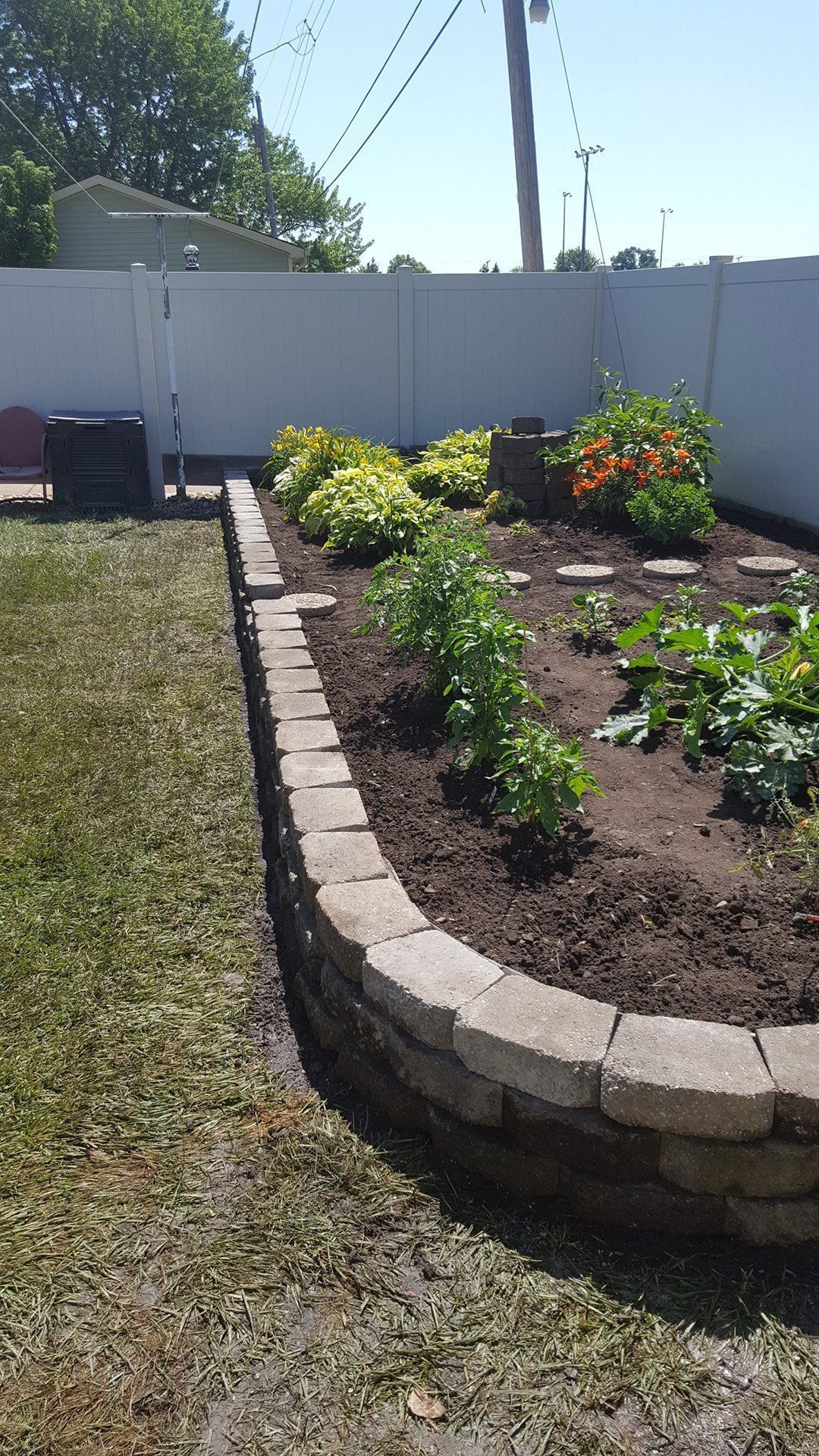 A curved garden bed with brick edging, filled with plants and dark soil, against a white fence.
