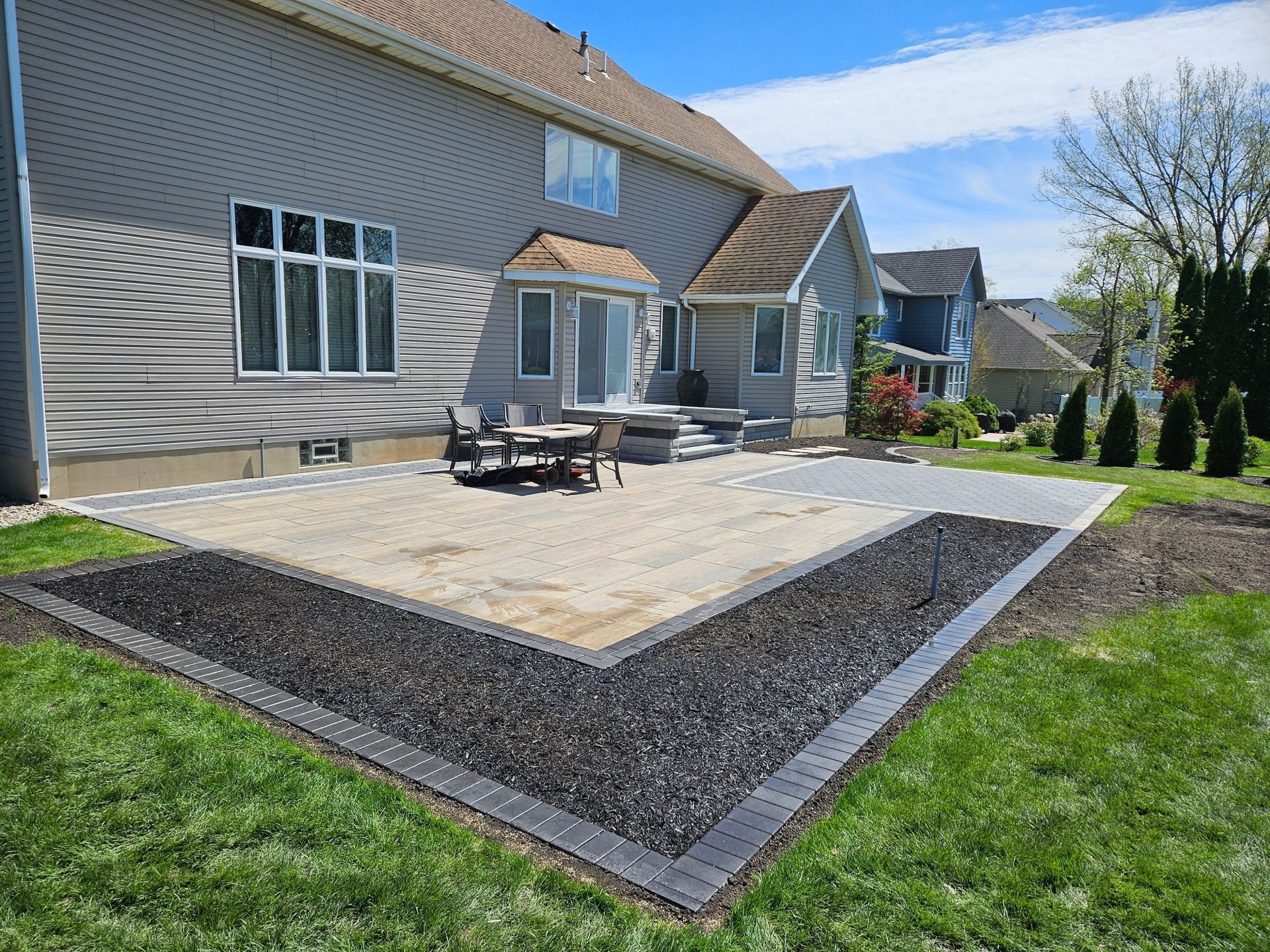 Backyard patio with gravel, dark border, and house in the background. Green grass surrounds it.