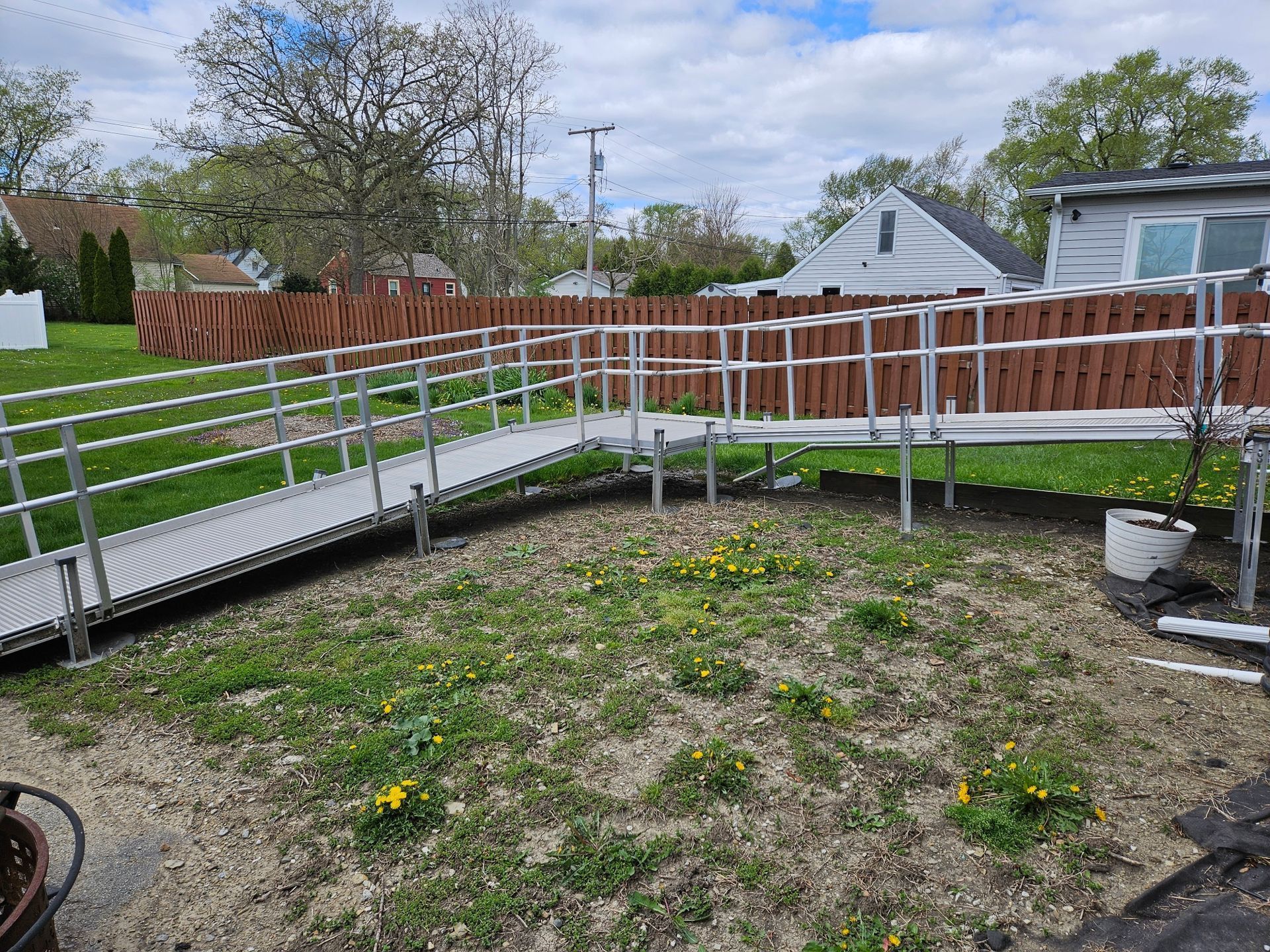 Exterior view of an aluminum wheelchair ramp in a yard with brown fence and houses in the background.